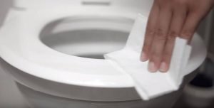 Hand wischt mit Toilettenpapier über einen weißen Toilettensitz - Reinigen des Toilettensitzes mit Desinfektionsliquid