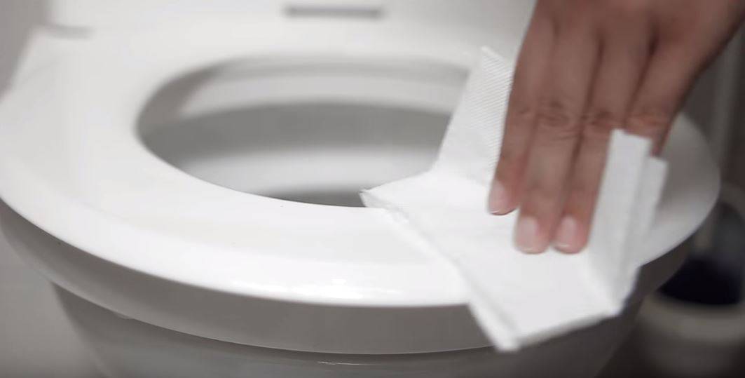 Hand wischt mit Toilettenpapier über einen weißen Toilettensitz - Reinigen des Toilettensitzes mit Schaumspender Surface Cleaner Toilet - Toilettensitzreiniger im günstigen CleanUp-Mietservice